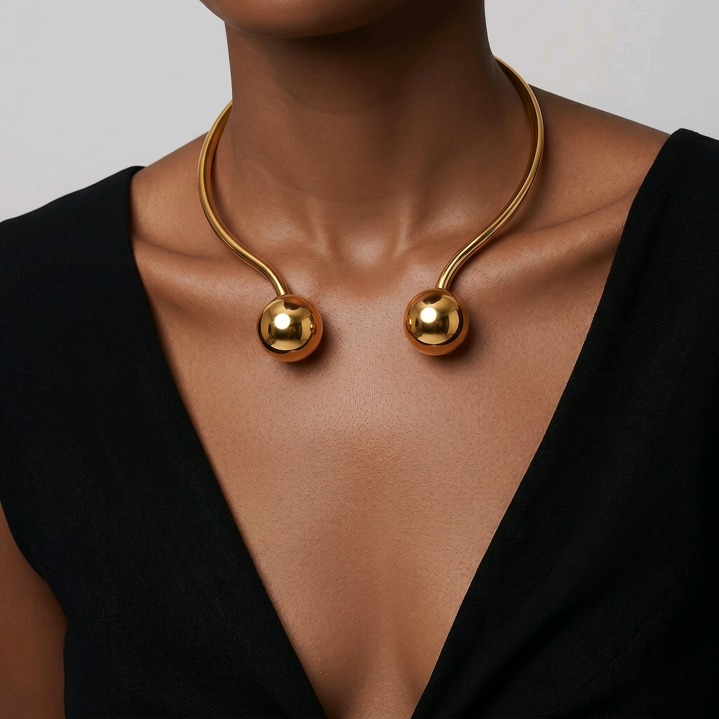 A close-up of a model wearing a brass-toned sculptural necklace with two spherical bead pendants.