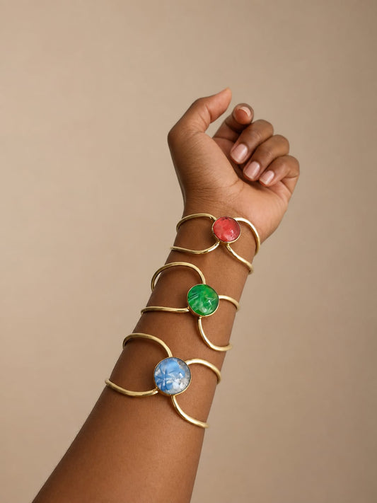 A person's forearm and hand wearing four overlapping brass-toned cuff bracelets, each with a round cabochon stone in red, green, and blue, against a plain beige background.