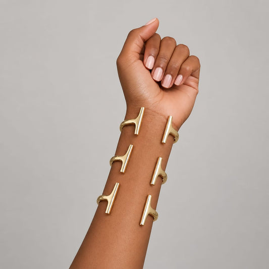 A close-up of a person's forearm and hand wearing six gold T-shaped cuff bracelets arranged vertically on the wrist against a plain light gray background.