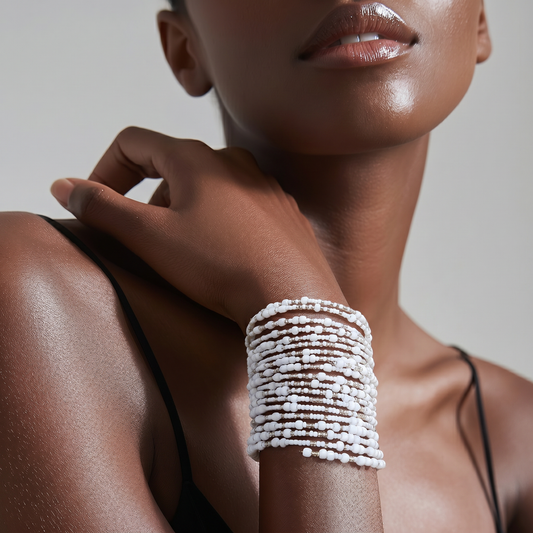 A close-up of a model wearing a thick, multi-strand cuff bracelet made of small white beads and silver-toned metal links, styled with a black spaghetti strap top against a neutral gray background.