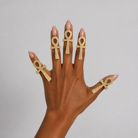 A hand with manicured nails wearing five textured Handmade brass ankh symbol rings, set against a solid light gray background.
