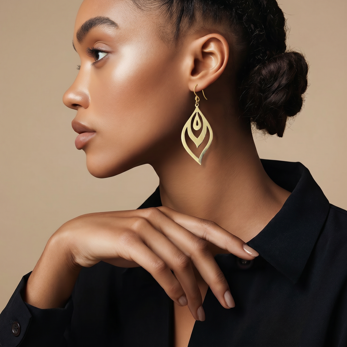 A woman in profile wearing a large gold leaf-shaped dangle earring, paired with a black collared shirt against a warm beige background.